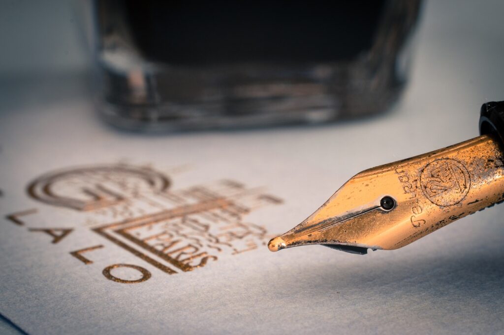 Złota stalówka pióra wiecznego, w tle kaligraficznie zapisane znaki i fragment kałamarza.